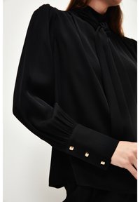 Blusa nera con colletto alto, maniche arricciate e tre bottoni quadrati dorati al polsino. Tessuto liscio con una leggera lucentezza.