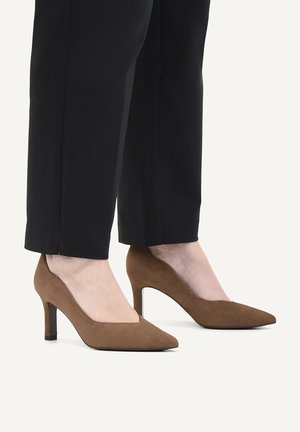 Brown suede pointed-toe high heels worn with black cropped pants on a light neutral background.