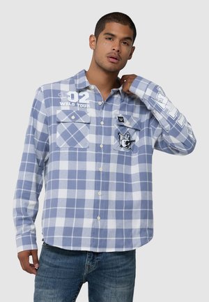Mann trägt blau-weiß kariertes Hemd mit Brusttaschen, grafischen Designs und blauen Jeans, posiert mit Hand nahe am Kragen.