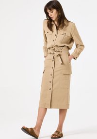 Beiges Utility-Kleid aus Baumwolle mit Hemdkragen, Knopfleiste, tailliertem Gürtel und zwei aufgesetzten Taschen, kombiniert mit braunen Riemchensandalen.