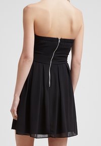Zwarte strapless jurk met een aansluitend lijfje, geplooide textuur en een uitlopende rok. De achterkant heeft een zichtbare zilveren rits en plooien.