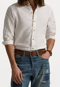 Bílá lněná košile s límečkem na knoflíky. Záhyby rukávů vyhrnuté, doplněné o poškozené modré džíny a hnědý kožený pásek. Na zápěstí kovové hodinky.