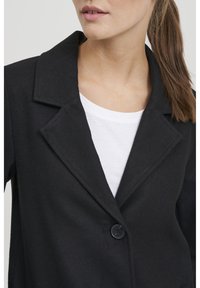 Cappotto in misto lana nero con colletto a scollatura, chiusura con un bottone e finitura testurizzata, indossato sopra una camicia bianca.