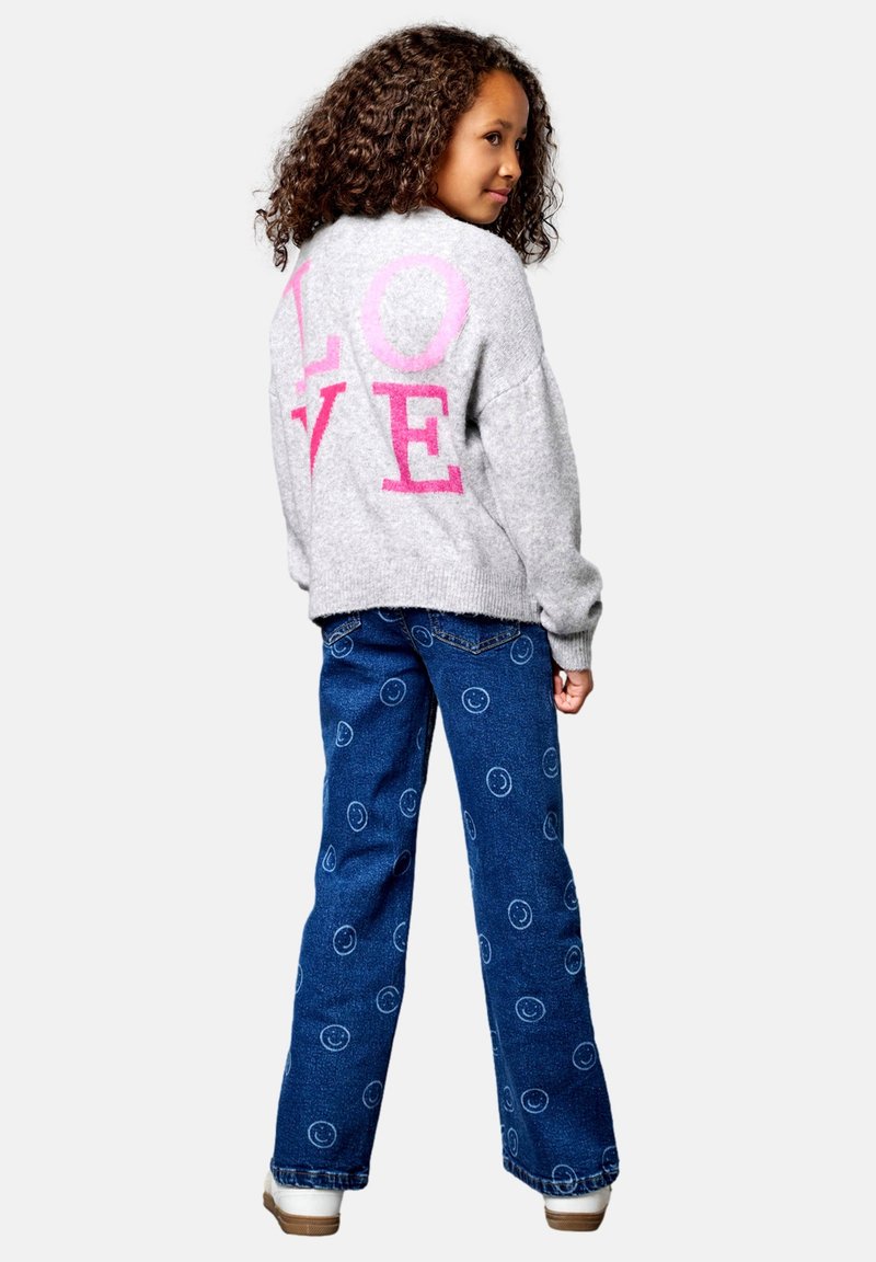 Fille aux cheveux bouclés portant un pull gris avec un texte rose "LOVE" dans le dos et un jean bleu avec des motifs de smileys blancs.