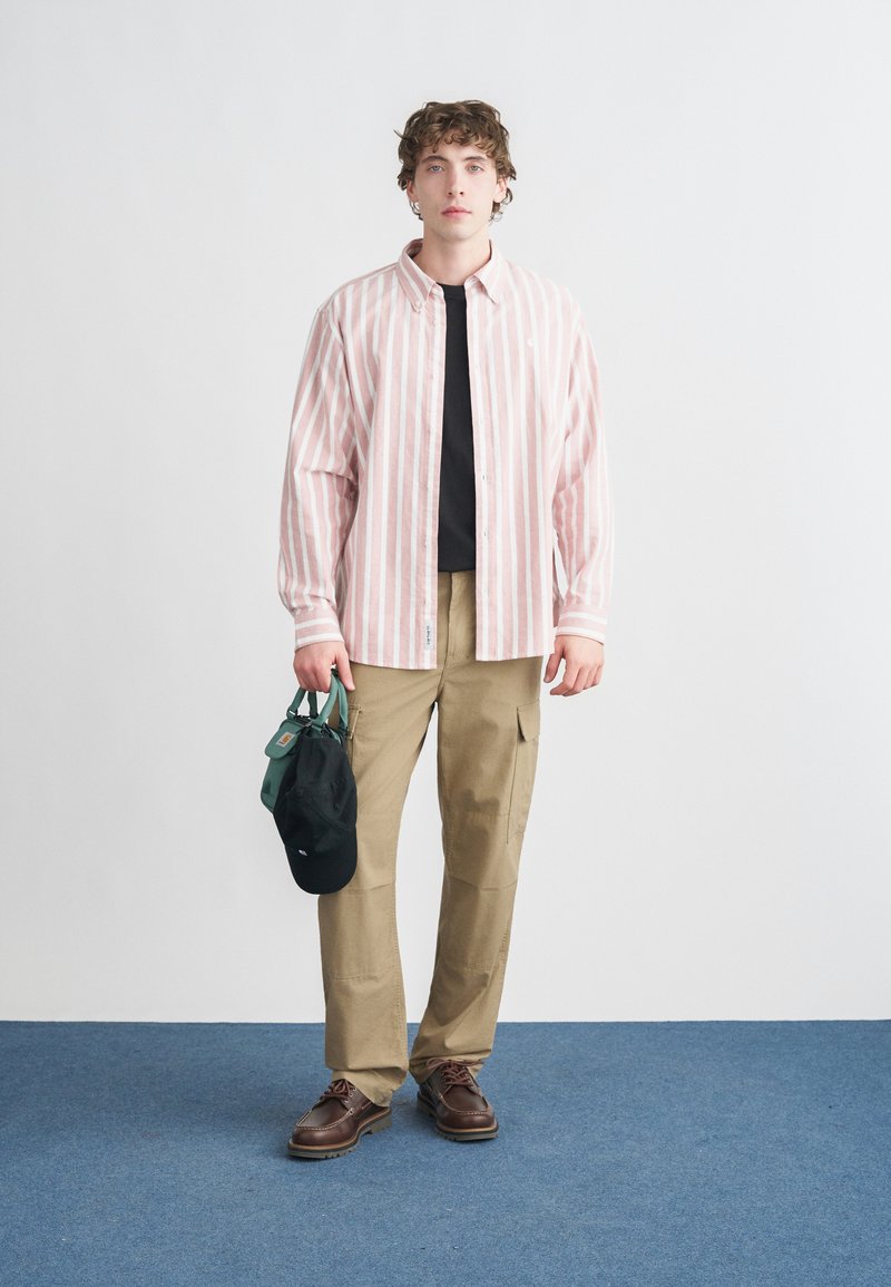 Chemise à rayures roses claires et blanches avec boutons, t-shirt noir en dessous, pantalon cargo beige, chaussures marron, tenant un sac vert et une casquette noire.