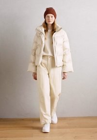 Chaqueta acolchada beige con cuello alto, suéter crema y pantalones anchos crema. Complementado con un gorro de punto marrón y zapatillas blancas.