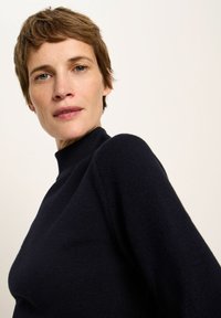 Frau mit kurzen braunen Haaren, die einen schwarzen, strukturierten Strickpullover mit hohem Kragen trägt und leicht nach unten sieht, vor einem schlichten, hellen Hintergrund.