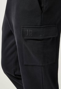 Černé kalhoty s boční kapsou na náklad s klopou a jemným vyraženým logem s písmeny a symbolem pí.