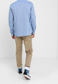 Homme portant une chemise à manches longues bleu clair, un pantalon beige retroussé, des chaussettes blanches et des baskets montantes bleues, marchant sur un sol blanc uni.