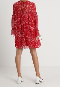 Robe florale rouge en tissu transparent, coupe décontractée, manches longues à bouffant, et détail en forme de trou de serrure au dos. Accentuée avec des chaussures à talons carrés blancs.