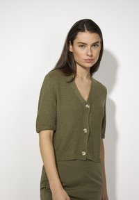 Kurzärmlige olivfarbene Strickjacke mit V-Ausschnitt, ausgestattet mit drei runden Knöpfen und gewebtem Strukturmuster, kombiniert mit olivfarbenen Hosen.