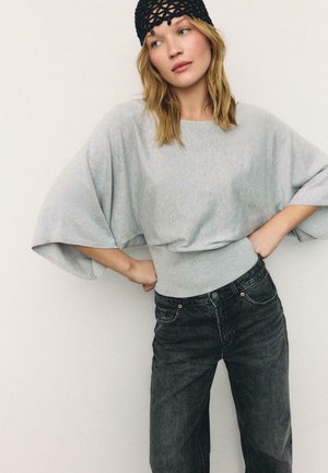 Žena nosící světle šedý top s širokými rukávy, černý tkaný klobouk a tmavé vysoké džíny, s rukama opřenýma v bok.