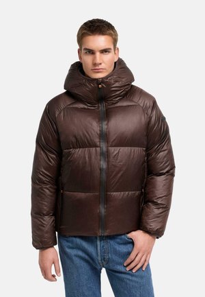 Ung mand iført en skinnende, mørkebrun dunjakke med høj krave og forlynlås, kombineret med blå jeans, stående mod en ensfarvet baggrund.
