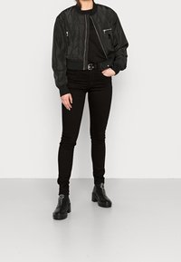 Mujer vestida con una chaqueta bomber negra, pantalones negros ajustados, botas negras de tobillo con cremalleras y una riñonera negra, de pie frente a un fondo liso.