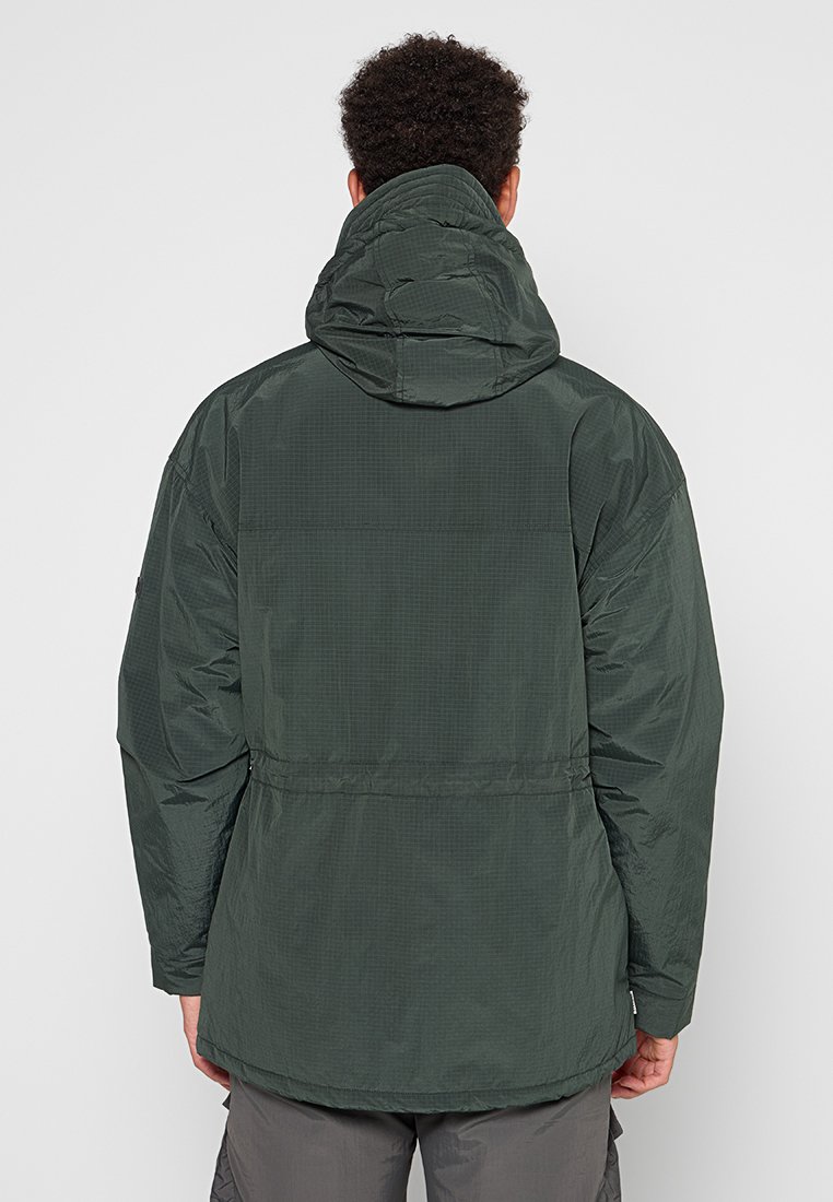 Mężczyzna w ciemnozielonej kurtce z kapturem, pokazany od tyłu, stojący na jednolitym jasnym tle.