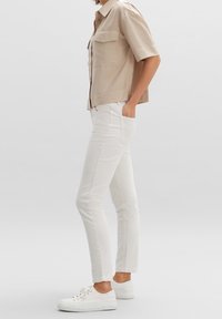 Vita slim-fit jeans med en slät yta och fem fickor, kombinerat med en kortärmad beige skjorta. Skorna är vita sneakers.