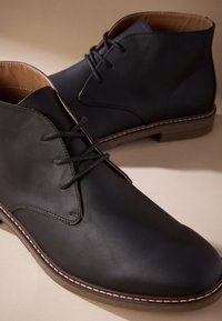 Čierne kožené členkové topánky s hladkou textúrou, zaoblenou špičkou a kontrastným bielym prešívaním okolo podrážky. Mávia šnurovanie.