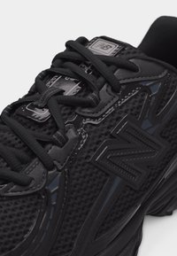 Black New Balance athletic shoe with mesh upper, thick sole, textured logo on the side, and black laces tied over the tongue.