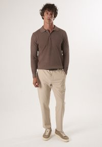 Bruine, gestructureerde polo met lange mouwen en een kraag, gecombineerd met beige broeken en lichtgekleurde sneakers, handen in de zakken.