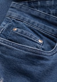 Primer plano de un bolsillo de jeans de mezclilla azul con costuras visibles y dos remaches metálicos en el pequeño bolsillo para monedas.