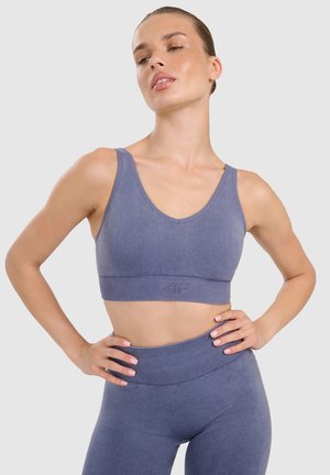 Kobieta pozuje z rękami na biodrach, ubrana w pasujący niebieski stanik sportowy i legginsy z wysokim stanem, na jednolitym jasnym tle.
