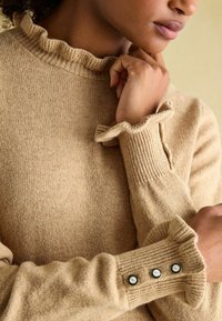 Beige gestrickter Pullover mit einem gerippten, gerafften Halsausschnitt und Bündchen. Verfügt über drei Knopfakzente am Bündchen. Weiche Textur, taillierte Passform.
