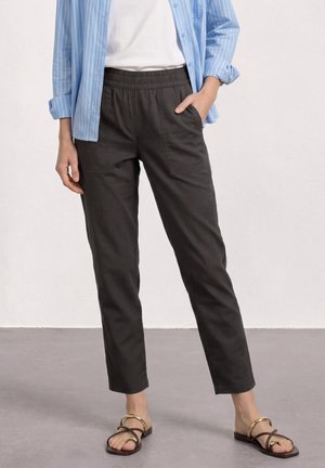 Personne portant un pantalon noir à taille élastique avec des poches avant, une chemise blanche, une chemise à rayures bleues à boutons, et des sandales à brides dorées.