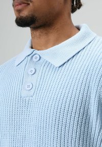 Close-up van een persoon die een lichtblauw gebreid poloshirt draagt met drie knopen en een kraag, waarbij de nek en de onderkant van het gezicht zichtbaar zijn.