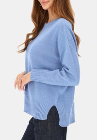 Kobieta w jasnoniebieskim swetrze z dzianiny z bocznymi rozcięciami i ciemnoniebieskich dżinsach, stojąca na tle jednolitego jasnego tła.