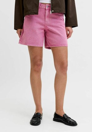 Žena v ružových džínových šortkách, čiernych mokasínach a tmavohnedej bunde s rukami pozdĺž tela, stojaca na bielom jednoduchom pozadí.