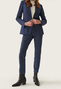 Námorně modrý přizpůsobený oblek z texturované látky, který zahrnuje blazer s jedním knoflíkem, slim-fit kalhoty a černé kotníkové boty.