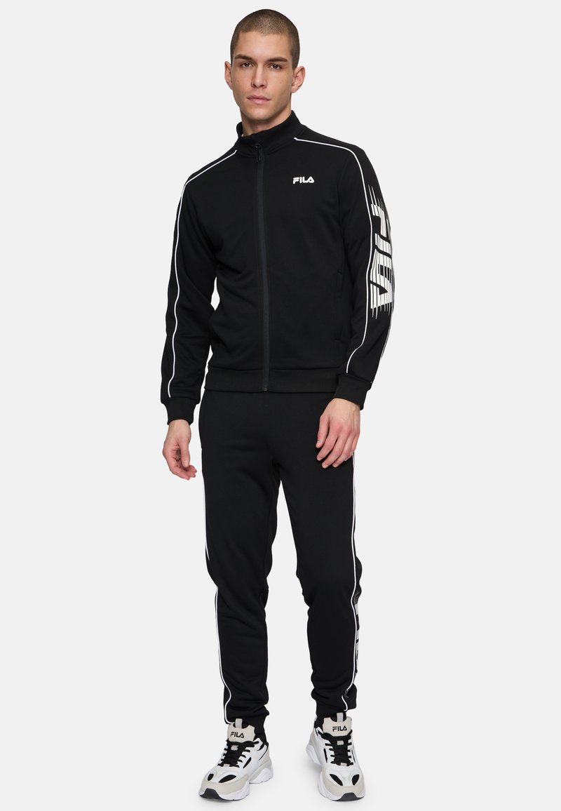 Svart träningsdräkt av mjukt material, med vita sidostreck, zip-up jacka och matchande byxor. Innehåller framträdande FILA-logotyp.