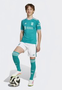 Έφηβος ποδοσφαιριστής σε τζαμένια φανέλα της Λίβερπουλ στέκεται με το ένα πόδι πάνω σε μια ασπρόμαυρη μπάλα ποδοσφαίρου μπροστά σε λευκό φόντο.