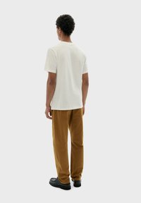 Witte katoenen T-shirt met korte mouwen, gecombineerd met bruine corduroy broek en zwarte schoenen; gezien van de achterkant.