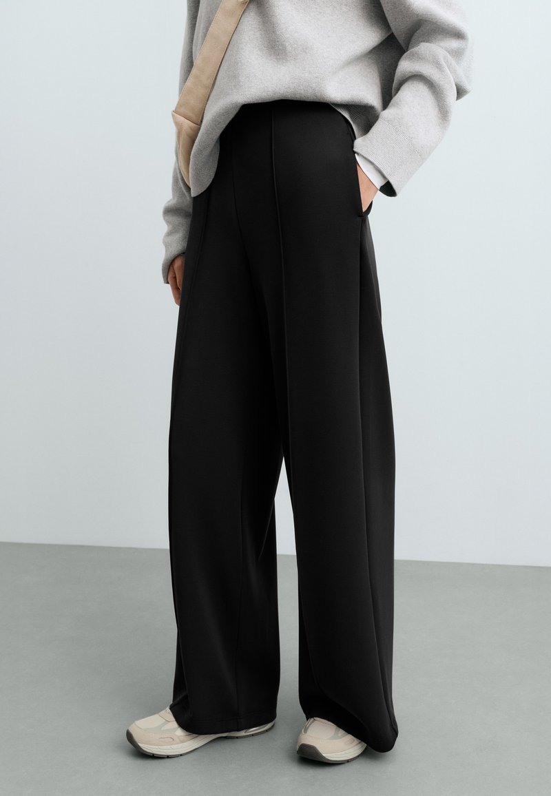 Personne portant un pantalon large noir, un pull gris, un sac beige en bandoulière et des baskets claires, debout sur un sol gris.