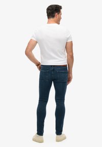 Jeans slim-fit de color azul oscuro con bolsillos traseros de parche, combinados con una camiseta blanca ajustada. Los zapatos son beige y de diseño sencillo.