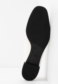 Chaussure en cuir blanc avec une semelle en caoutchouc noire. Le talon est fin et a une finition lisse, avec un logo en relief discret.