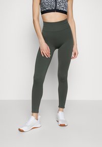 Legginsy w kolorze ciemnozielonym, z wysokim pasem i fakturowanym wzorem, połączone z krótkim topem w wzory. Białe buty sportowe z pomarańczowymi akcentami.