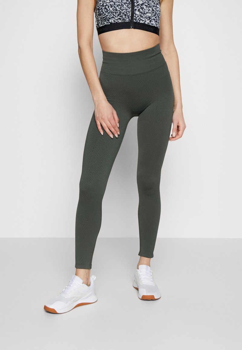 Legginsy w kolorze ciemnozielonym, z wysokim pasem i fakturowanym wzorem, połączone z krótkim topem w wzory. Białe buty sportowe z pomarańczowymi akcentami.