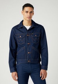 Chaqueta de mezclilla en azul oscuro con costuras de cobre, que presenta un cuello puntiagudo, cierre frontal de botones y dos bolsillos en el pecho con solapas.