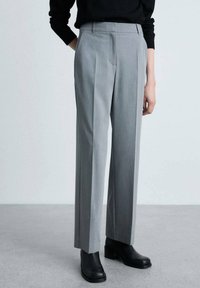 Pantalons gris ajustés avec une texture lisse, dotés de plis avant, d'une coupe droit et d'une taille mi-haute. Portés avec des bottines noires.