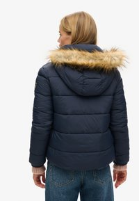 Doudoune navy avec capuche ornée d'une fausse fourrure. Le design présente des coutures horizontales et des poignets élastiques, associés à des manches en tricot léger.