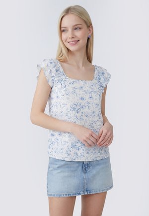Młoda kobieta o blond włosach, ubrana w białą kwiecistą bluzkę z falbankami na rękawach i jasnoniebieską spódnicę jeansową, uśmiechająca się i patrząca w bok.