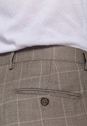 Primo piano di pantaloni a quadri marroni con una tasca posteriore abbottonata, indossati con una camicia bianca testurizzata infilata dentro.