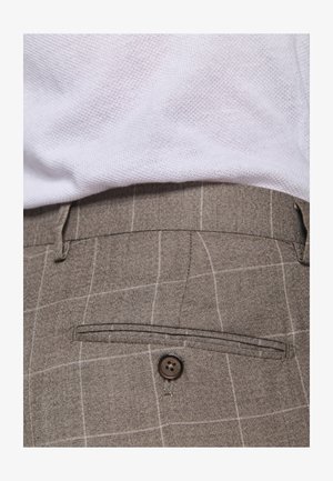 Primo piano di pantaloni a quadri marroni con una tasca posteriore abbottonata, indossati con una camicia bianca testurizzata infilata dentro.