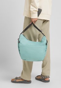 Türkisfarbene Schultertasche mit gepolstertem Design, doppelten schwarzen Trägern und Reißverschluss. Getragen von einer Person in beigen Hosen und Sandalen.