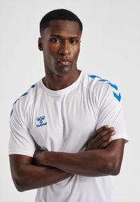 Wit sportshirt met blauwe accenten op de schouders, met een ronde hals en korte mouwen. Logo gedrukt op de linker borstzijde.