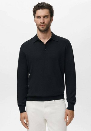 Uomo con capelli ricci corti che indossa una polo nera a maniche lunghe e pantaloni bianchi su uno sfondo chiaro uniforme.