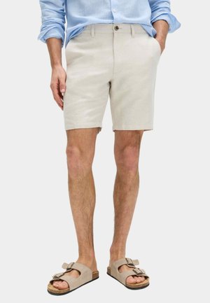 Pantalones cortos beige claro hechos de un tejido texturizado, con cierre de botón y bolsillos laterales. Combinados con sandalias marrón claro.