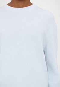 Jasnoniebieski dziergany sweter w prążki z okrągłym dekoltem, noszony przez osobę o średnio-ciemnym kolorze skóry, na jednolitym tle.